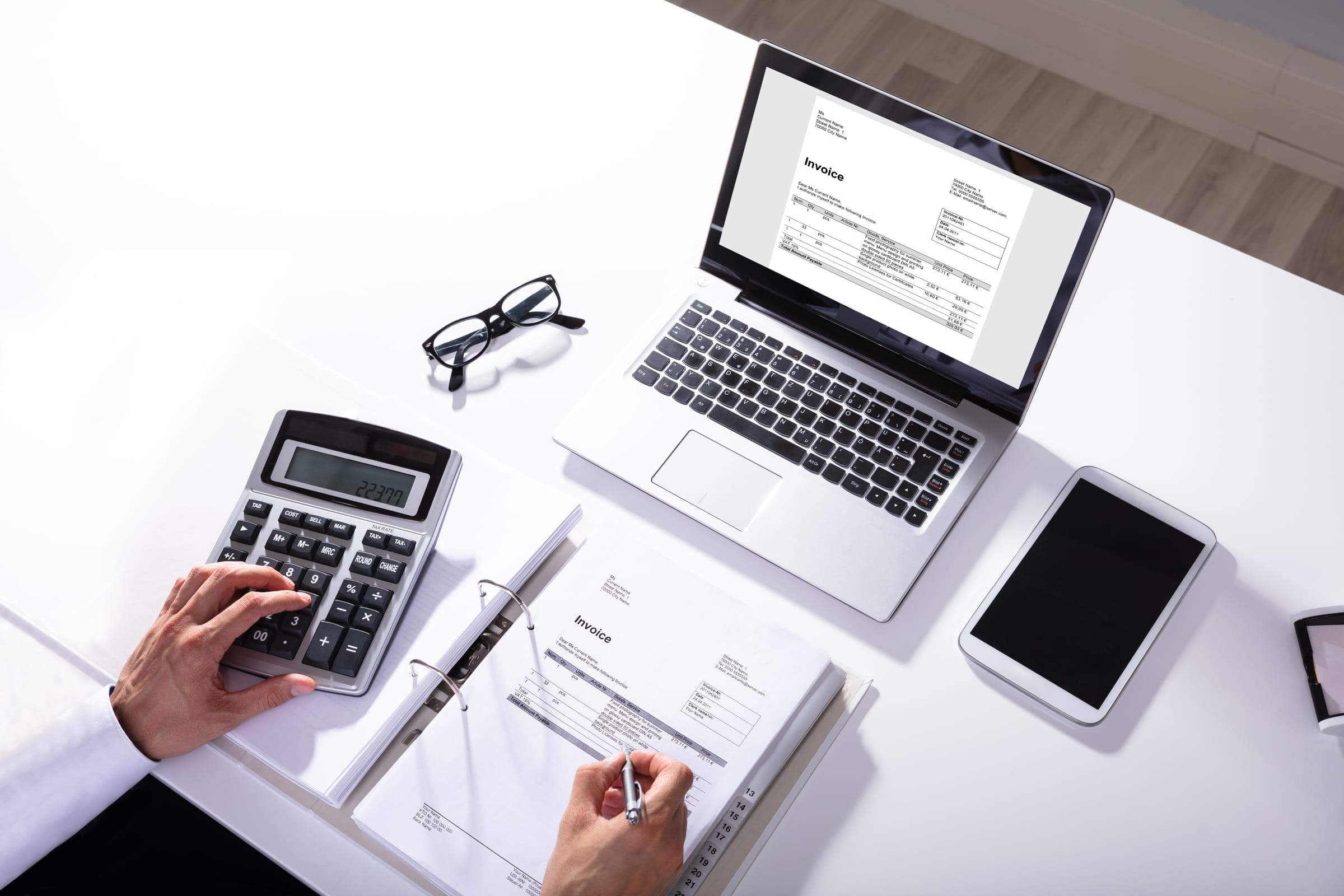 Hands using a desk calculator beside a laptop and printed invoices